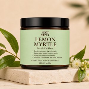 A jar of Secret Honey Lemon Myrtle Tallow Cream sits on a light stone slab, surrounded by green leaves and small white flowers against a warm beige background.