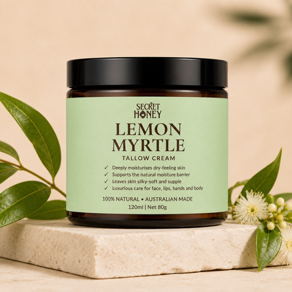 A jar of Secret Honey Lemon Myrtle Tallow Cream sits on a light stone slab, surrounded by green leaves and small white flowers against a warm beige background.