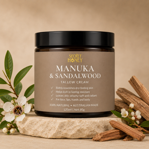 A jar of Secret Honey Manuka & Sandalwood Tallow Cream sits on a stone slab with manuka leaves, white flowers, and sandalwood bark around it, against a warm beige background.