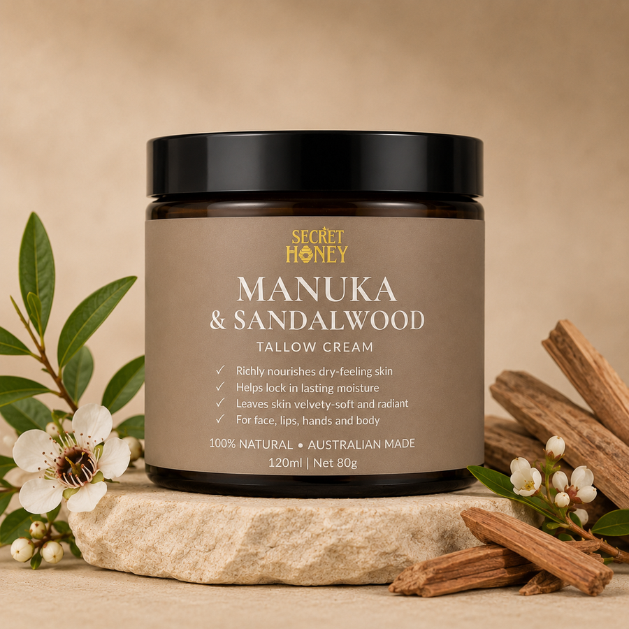 A jar of Secret Honey Manuka & Sandalwood Tallow Cream sits on a stone slab with manuka leaves, white flowers, and sandalwood bark around it, against a warm beige background.