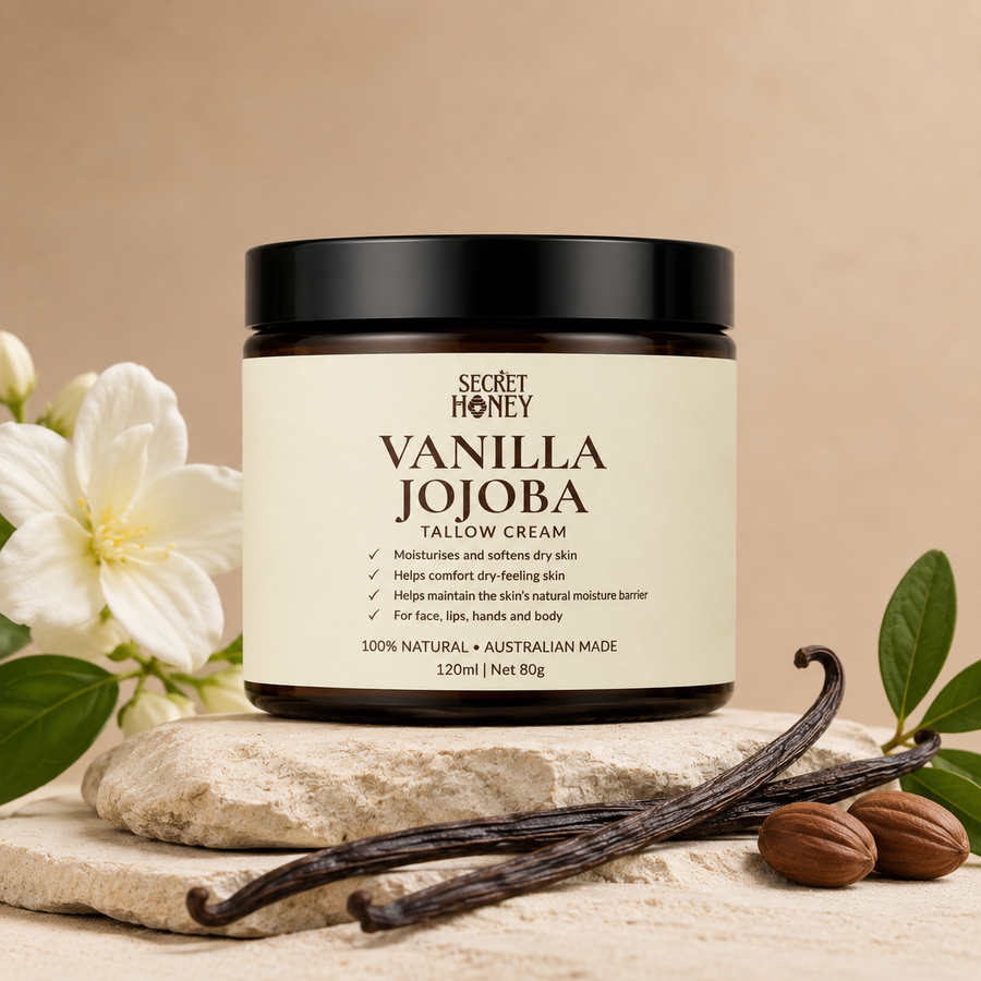 A jar of Secret Honey Vanilla Jojoba Tallow Cream is displayed on a stone slab with vanilla beans, almonds, and white flowers against a warm beige background.